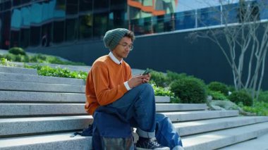 Kentsel basamaklarda oturan rahat bir ortamda mesaj atarken akıllı telefon ekranına odaklanan sıradan bir genç adam..