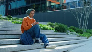 Şehir basamaklarında oturan şık genç adam, sakin ve modern bir açık hava ortamında akıllı telefonundaki çevrimiçi içeriğe göz atıyor..