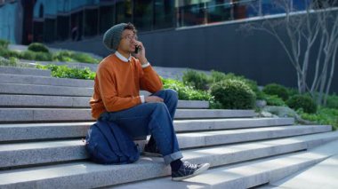 Gündelik giysiler giyen genç bir öğrenci ve bereli dışarıda merdivenlerde oturuyor ve akıllı telefondan konuşuyor, rahat ve odaklanmış..