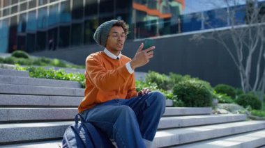 Sıradan genç adam şehrin merdivenlerinde oturuyor, elinde video görüşmesi için akıllı bir telefon ya da modern şehir ortamında selfie çekiyor..