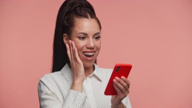 Akıllı telefona bakarken heyecanla gülümseyen Afro-Amerikalı kadın, yüz yüze, pembe arka planda soyutlanmış..