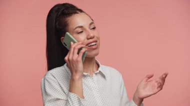 Akıllı telefondan konuşan neşeli bir kadın, sohbetin ortasında, gülümseyen ve el kol hareketi yapan, pembe arka planda soyutlanmış..