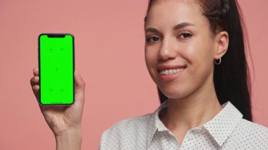 Kendine güveni tam bir kadın açık yeşil ekranlı akıllı telefonu elinde tutuyor. Model veya uygulama ekranı için, gülümsüyor, pembe arka planda soyutlanıyor..
