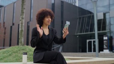Profesyonel bir kadın modern ofis binasının dışında oturuyor, elinde akıllı telefon ve bir video görüşmesi sırasında el sallıyor, gülümsüyor ve nişanlanıyor..
