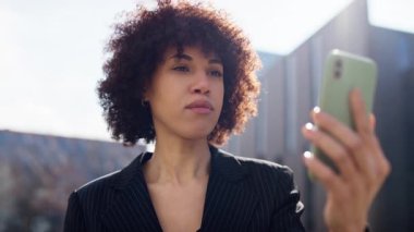 Gülümseyen kadın, akıllı telefondaki video görüşmesini bitirirken onaylayıp el sallıyor. Modern ofis binasının yanında duruyor..