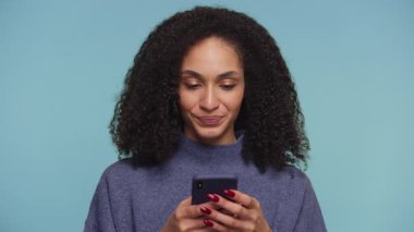 Kutlamada gülümseyen ve yumruklarını kaldıran heyecanlı genç kadın mavi arka planda akıllı telefonu tutuyor..