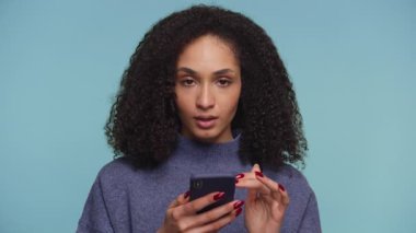 Genç bir kadın, akıllı telefona bakarken, mavi arka planda beklenmedik ya da üzücü haberlere tepki verirken ağzı inançsızlıkla kapatıyor..