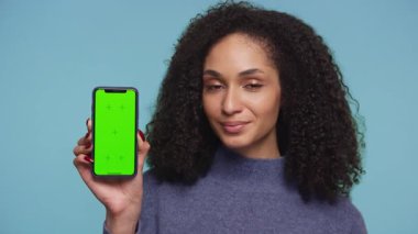 Kendine güvenen genç bir kadın gülümseyerek başını sallıyor, elinde yeşil ekranlı akıllı bir telefon tutuyor, mavi arka plana karşı aynı fikirde ya da memnuniyeti gösteriyor..