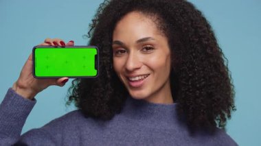 Neşeli genç bir kadın göz kırpıyor, el hareketi yapıyor ve mavi arka planda yeşil ekranlı akıllı telefonu tutuyor..