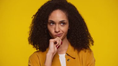 Hardal rengi ceketli kıvırcık saçlı kadın, elini çenesine uzatmış, derin düşüncelere dalmış, sarı arka planda izole edilmiş bir seçim ya da fikri düşünürken..