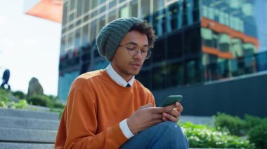 Sıradan, gözlüklü ve bereli bir genç adam merdivenlerde oturmuş, akıllı telefonuna bir mesaj yazarken,.