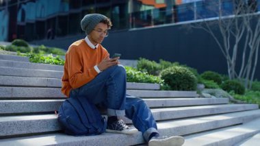 Şaşırmış ve endişeli genç adam dışarıda merdivenlerde oturuyor, şok içinde ağzı örtülmüş telefonundan beklenmedik kötü haberler okuyor..