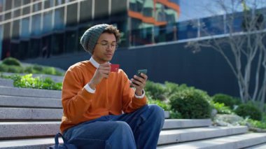 Günlük giysiler içinde şehir merdivenlerinde oturan, kredi kartı bilgilerini online alışveriş için akıllı telefona kaydeden odaklanmış genç bir adam..