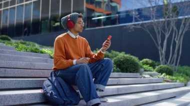 Kırmızı kulaklık takan ve günlük kıyafet giyen gülümseyen genç bir adam bir video görüşmesine katılıyor, akıllı telefondan selamlıyor ve dışarıda otururken konuşuyor..