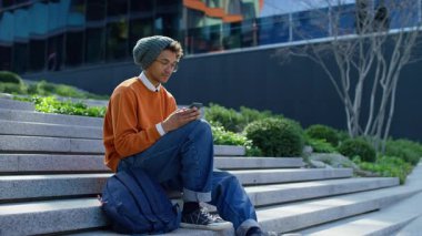 Neşeli genç adam dışarıda merdivenlerde oturuyor, sevinçle yumruğunu kaldırıyor ve gülümsüyor. Bu arada akıllı telefondaki büyük haber veya başarıya tepki veriyor..