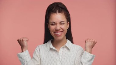 Genç bir kadın, yumuşak pembe bir arka planda, neşeli ve heyecanlı bir şekilde gülümsüyor..