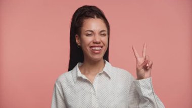 Puantiyeli neşeli genç bir kadın pembe arka planda parmaklarıyla barış işareti yapıyor..