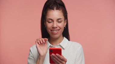 Genç kadın kameraya gülümserken akıllı telefona tıklıyor. Gurur, başarı ve dijital başarı duygusu ile pembe arka planda izole edilmiş.