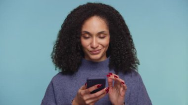 Kıvırcık saçlı kadın telefonuna neşeli ve belirsiz bir ifadeyle bakıyor. İnternetten alışveriş yaparken bir şey alıp almayacağını düşünüyor. Mavi arka planda soyutlanmış..
