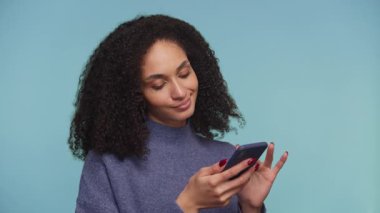 Genç bir kadın akıllı telefonuna bakarken nazikçe gülümsüyor, mavi arka planda izole edilmiş online bir alışveriş uygulamasına bakarken ürünlere olumlu tepki veriyor..