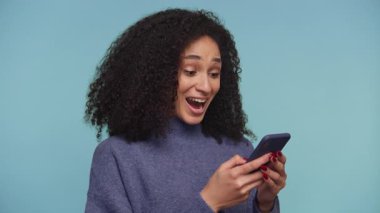 Telefonuna bakıp gülümseyen neşeli kadın kişisel başarısını ya da internetteki heyecan verici haberini kutluyor, mavi arka planda izole edilmiş..