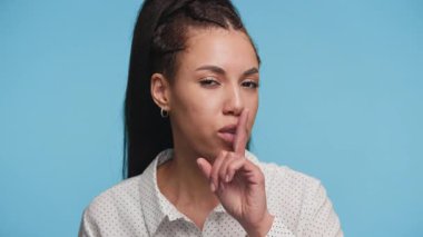 Dudakları üzerinde parmağı olan genç bir kadın, temiz, açık mavi bir arkaplanda sessizlik ya da gizlilik işareti yapıyor..