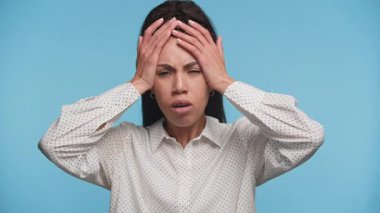 Hayal kırıklığı ya da zihinsel stres gösteren genç bir kadın, iki eliyle mavi bir arka plana karşı başını tutuyor..