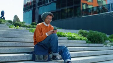 Modern binanın yanındaki merdivenlerde oturan şık genç adam, akıllı telefonuna bir mesaj yazmaya odaklanmış..