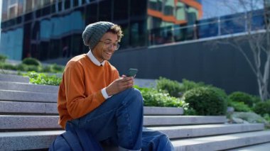 Gülümseyen üniversite öğrencisi dışarıda merdivenlerde oturuyor, sevinçle olumlu haberler okuyor ya da bir kampüs binasının yakınındaki akıllı telefonundan mesaj okuyor..