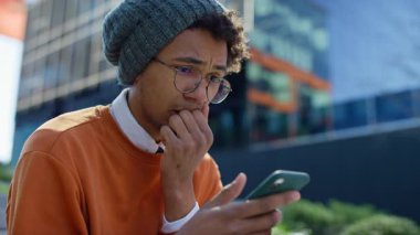 Gündelik giysiler içindeki endişeli üniversite öğrencisi, modern bir binanın dışındaki akıllı telefonundan hatalı sınav sonuçları aldıktan sonra stresli görünüyor..