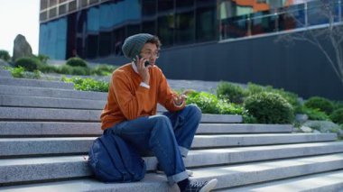 Dışarıdaki merdivenlerde oturan genç bir öğrenci, telefondan kulağa, modern bir binanın yakınında gündelik ya da biraz karışık bir sohbete dalmış..