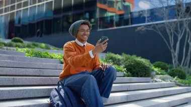 Kampüs basamaklarında oturan gülümseyen üniversite öğrencisi rahat ve güneşli bir ortamda akıllı telefonuyla video görüşmesinin tadını çıkarıyor..