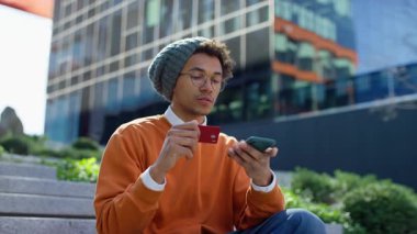Üniversite öğrencisi açık havada elinde kredi kartıyla oturuyor. İnternetten ödeme yapıyor ya da kampüs merdivenlerinden alışveriş yapıyor..
