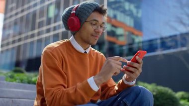 Kampüs basamaklarında oturan genç bir üniversite öğrencisi, kulaklık takıyor ve rahat bir ortamda akıllı telefondan internet içeriğini tarıyor..