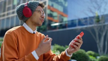 Üniversite öğrencisi dışarıda merdivenlerde oturuyor, kulaklık takıyor ve akıllı telefondan bir video görüşmesi yapıyor, konuşurken işaret veriyor..