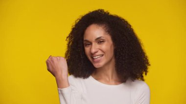 Beyaz bluzlu gülümseyen genç kadın, canlı sarı bir arka plana karşı neşe ve başarısını ifade eden bir kutlama hareketi yapıyor..