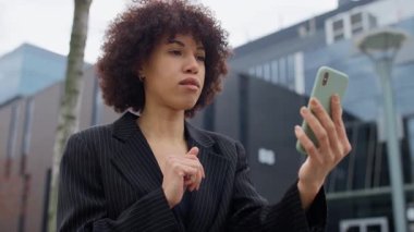 İnce çizgili takım elbiseli profesyonel bir kadın akıllı telefonuyla bir video görüşmesi yapıyor, modern bir ofis binasının önünde duruyor..