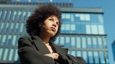 Kollarını kavuşturmuş kararlı bir kadın, mavi gökyüzünün altında cam gökdelenlerle çevrili bir iş bölgesinde güvenle dikiliyor..