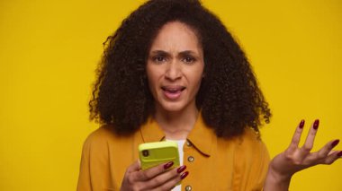 Kafası karışmış ve rahatsız olmuş genç bir kadın, sarı arka plandaki beklenmedik ya da kötü haberlere tepki göstererek akıllı telefonuna hayal kırıklığıyla bakar..