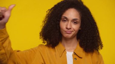 Memnun olmayan genç bir kadın ciddi bir ifadeyle baş parmak göstererek, canlı sarı bir arka plana karşı memnuniyetsizliğini ya da memnuniyetsizliğini dile getiriyor.