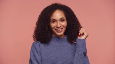 Mavi kazaklı neşeli genç bir kadın kameraya sıcak bir şekilde gülümsüyor, stüdyo ışıklarında yumuşak pembe bir zemin üzerinde duruyor..