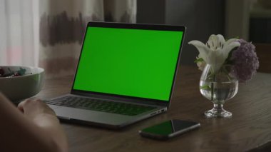 Ahşap bir masada yeşil ekranlı modern bir laptop, akıllı bir telefonun yanında, vazoda çiçekler, ve bir kase kiraz..