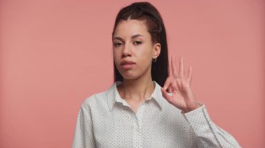 Sakin bir yüz ifadesi olan genç bir kadın iyi bir el hareketi yapıyor. Yumuşak pembe bir arka plana karşı güvenle duruyor..