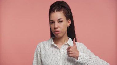 Beyaz tişörtlü genç bir kadın, pembe arka planda duran, kompleks bir ifadeyle baş parmağını kaldırıyor..