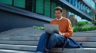 Gözlüklü genç adam şok içinde bilgisayar ekranına bakarken, modern bir ofis binasının önünde oturuyor..