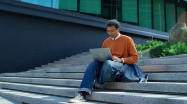 Genç bir öğrenci elinde dizüstü bilgisayarla dışarıda oturuyor, kampüsteki modern bir akademik binanın yanında çalışıyor ya da okuyor..