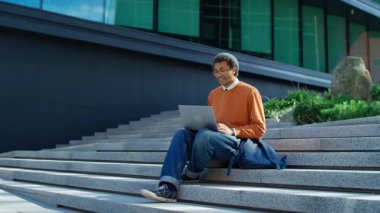 Kampüs basamaklarında oturan genç öğrenci video görüşmesi için dizüstü bilgisayar kullanıyor, modern ortamlarda iletişim kuruyor ya da ders çalışıyor..