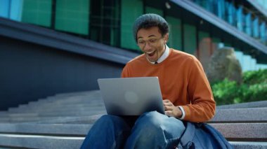 Heyecanlı genç adam dışarıda basamaklarda oturur, laptoptan olumlu haberler okur, ağzı neşeli inançsızlıkla ve sürprizle kaplıdır..