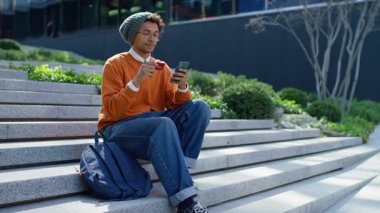 Merdivenlerde oturan genç adam alışveriş yapmak için akıllı telefon ve kredi kartı kullanıyor ya da internetten ödeme yapıyor, rahat bir açık hava anının tadını çıkarıyor..