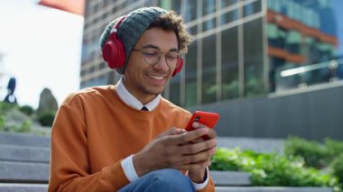 Dışarıda oturan mutlu genç adam, telefonuna mesaj atıyor ve kulaklık takıp gülerken komik bir mesaja gülüyor..